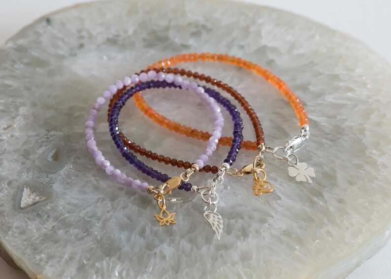 Peut inclure: Trois bracelets en perles avec des breloques. Les bracelets sont superpos&eacute;s. Le bracelet du haut est orange, le bracelet du milieu est violet et le bracelet du bas est violet clair. Les breloques sont une fleur de lotus dor&eacute;e, une aile argent&eacute;e et un tr&egrave;fle &agrave; quatre feuilles argent&eacute;.
