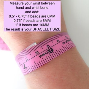 Op de afbeelding: Een roze meetlint gewikkeld om een pols met instructies om de pols te meten tussen de hand en het polsbot en 0,5 inch toe te voegen als de kralen 6 mm zijn, 0,75 inch als de kralen 8 mm zijn of 1 inch als de kralen 10 mm zijn. Het resultaat is de armbandmaat.