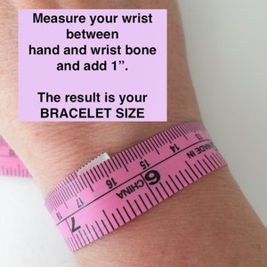 Op de afbeelding: Een roze meetlint gewikkeld om een pols om te laten zien hoe je de maat van een armband meet. Het lint heeft de tekst "MADE IN" en "6 CHINA". De tekst op de afbeelding luidt "Meet je pols tussen hand en polsbeen en voeg 1 toe". Het resultaat is je ARMBANDMAAT.