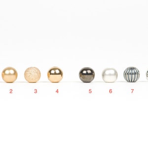May include: Eight different round beads in a row. The beads are gold, silver, white, and black. The beads have different textures, including smooth, textured, and ribbed.
