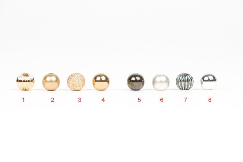 May include: Eight different round beads in a row. The beads are gold, silver, white, and black. The beads have different textures, including smooth, textured, and ribbed.