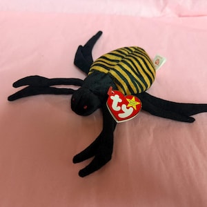May include: A black plush toy spider with a yellow and black striped body. The spider has long, black legs and red eyes. A red heart-shaped tag with white text and a gold star is attached to the toy.