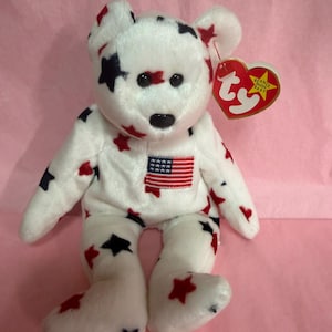 May include: A white plush bear with red and navy blue star patterns and an American flag patch. The bear has a black nose and eyes, and a red heart-shaped tag that says "Beanie Baby".