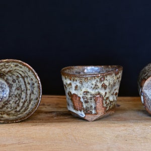 May include: Three ceramic cups with a speckled brown and white glaze. The cups are arranged in a row on a wooden surface.