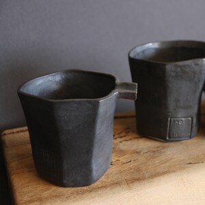 May include: Three black ceramic cups with a geometric design. The cups are sitting on a wooden surface.