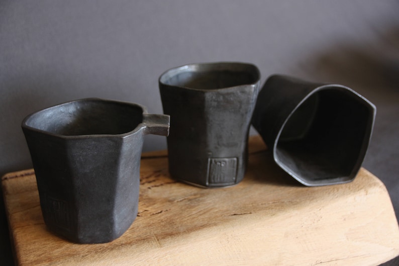 May include: Three black ceramic cups with a geometric design. The cups are sitting on a wooden surface.