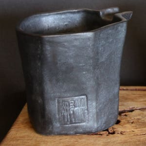 May include: A black ceramic pot with a unique, angular shape and a raised, rectangular design on the side. The pot has a slightly uneven surface and a rough texture.