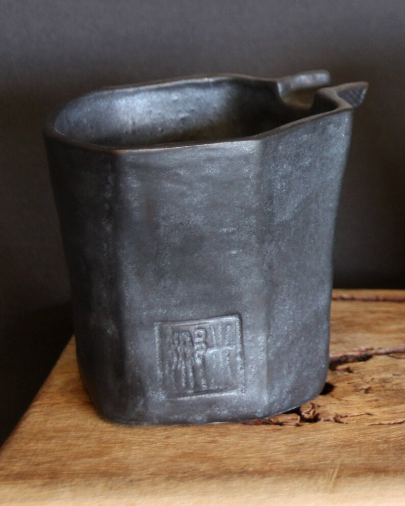 May include: A black ceramic pot with a unique, angular shape and a raised, rectangular design on the side. The pot has a slightly uneven surface and a rough texture.