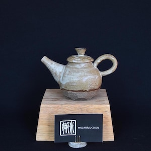 Può includere: Un piccolo teiera in ceramica bianca con una glassa marrone e un coperchio. La teiera è appoggiata su un supporto in legno con un biglietto da visita che recita "Minna Graham Ceramics".