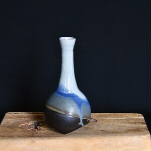 May include: A small, ceramic vase with a rounded base and a narrow neck. The vase is glazed in shades of blue, grey, and black.