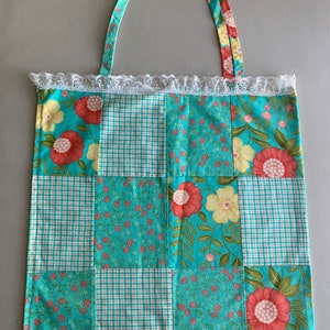 以下が含まれることがあります： 花柄と格子柄のターコイズブルーのパッチワークトートバッグ。バッグは白いレースのトリムと2つのファブリックハンドルが付いています。