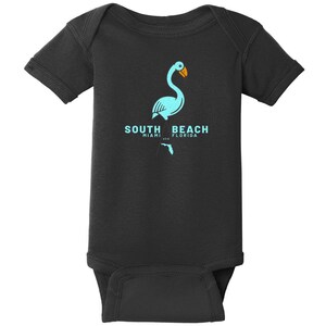 May include: A black short-sleeve bodysuit featuring a light blue graphic of a flamingo with an orange beak. Below the flamingo, the text reads SOUTH BEACH MIAMI FLORIDA USA, accompanied by a small light blue silhouette of the state of Florida.