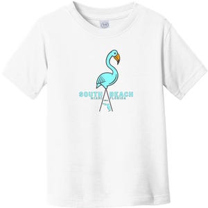 May include: A white short-sleeve t-shirt featuring a light blue flamingo graphic with an orange beak. Below the flamingo, the text reads SOUTH BEACH MIAMI FLORIDA USA, accompanied by a small outline of the state of Florida in light blue.