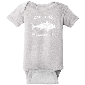 May include: A heather gray short-sleeve baby bodysuit featuring a white graphic of a fish in the center. Above the fish, the text reads CAPE COD, and below the fish, the text reads MASSACHUSETTS. The garment has a crew neckline and snap closures.