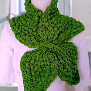 May include: A vibrant green knitted scarf with a leaf-like design. The scarf is made of textured yarn and is draped over a white top. The scarf's intricate pattern and bright color make it a striking accessory.