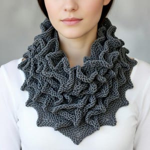 以下が含まれることがあります： グレーのフリルデザインのニットカウル。柔らかく、質感のある糸で作られたスカーフで、下部は三角形です。カウルは首の周りに着用します。
