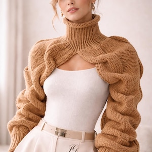 May include: A woman wearing a tan-colored, chunky knit bolero sweater with a high turtleneck collar. The sweater has a unique design with open front and sleeves. The model is also wearing a white tank top, cream-colored pants, and a belt. The brand name "JEN GIEZEN" is visible.