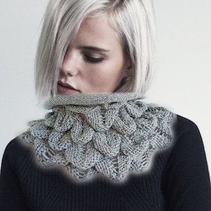 May include: A gray knitted cowl with a unique, textured design. The cowl is made of a soft, chunky yarn and has a wide, open neckline.