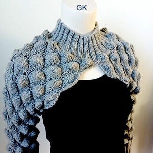 Op de afbeelding: Grijze gebreide shrug met een textuur, bubbelachtig patroon. De shrug heeft een hoge hals en lange mouwen. Het label "GK" is zichtbaar op de nek van het mannequin.