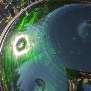 May include: A close-up shot of a glass bowl with a green and blue swirled design. The bowl has a reflective surface with a bright white light reflecting off the surface. The rim of the bowl is silver.