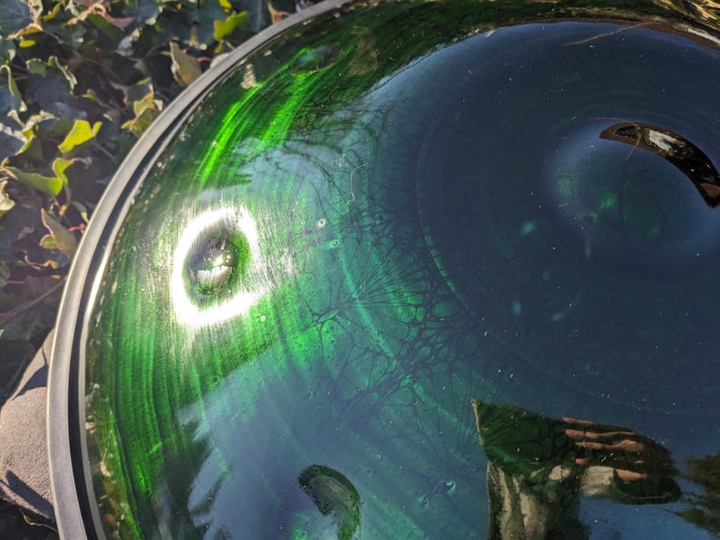 May include: A close-up shot of a glass bowl with a green and blue swirled design. The bowl has a reflective surface with a bright white light reflecting off the surface. The rim of the bowl is silver.