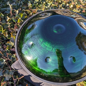 May include: A green and black handpan drum with a reflective surface. The instrument has a circular shape with several dimples and a dark rim. The handpan is resting on a stone surface, surrounded by green foliage.