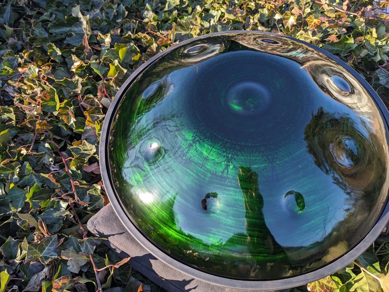 May include: A green and black handpan drum with a reflective surface. The instrument has a circular shape with several dimples and a dark rim. The handpan is resting on a stone surface, surrounded by green foliage.