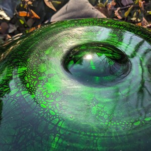 May include: Close-up of a green glass bowl with a textured surface and a central, rounded indentation. The bowl's surface features a dark, web-like pattern, and the glass reflects light, creating highlights.