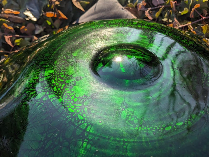May include: Close-up of a green glass bowl with a textured surface and a central, rounded indentation. The bowl's surface features a dark, web-like pattern, and the glass reflects light, creating highlights.