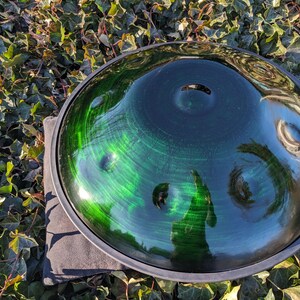 May include: A handpan, a steel percussion instrument, with a deep green and blue finish. The instrument has a circular shape with several dimples and a dark rim. The handpan is resting on a gray surface, surrounded by green foliage.