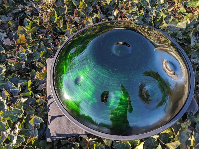 May include: A handpan, a steel percussion instrument, with a deep green and blue finish. The instrument has a circular shape with several dimples and a dark rim. The handpan is resting on a gray surface, surrounded by green foliage.