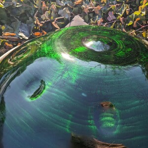 May include: A close-up shot of a green glass bowl with a swirling pattern. The bowl has a dark green hue with lighter green accents, creating a textured appearance. The rim is a silver metal. The background is blurred with green and brown foliage.