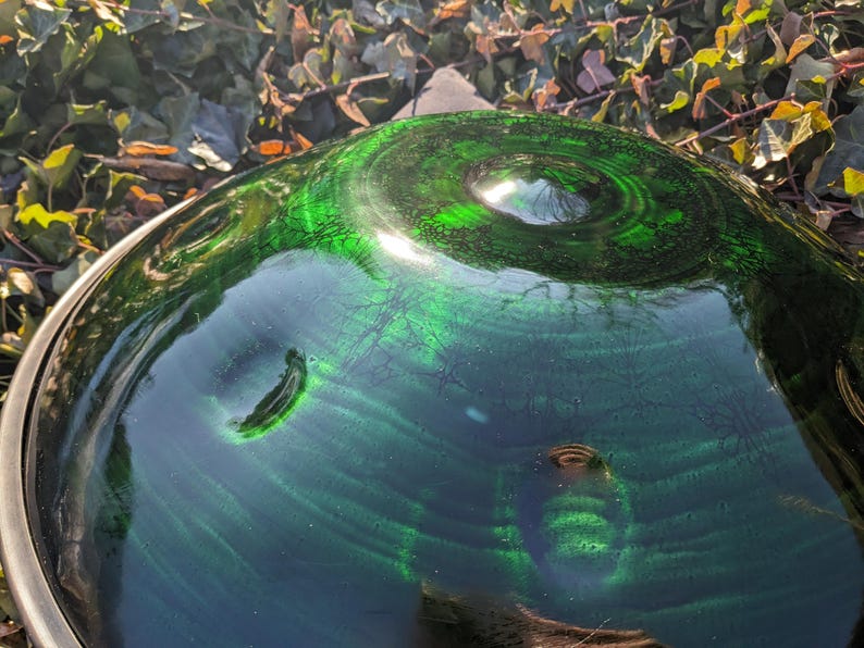 May include: A close-up shot of a green glass bowl with a swirling pattern. The bowl has a dark green hue with lighter green accents, creating a textured appearance. The rim is a silver metal. The background is blurred with green and brown foliage.