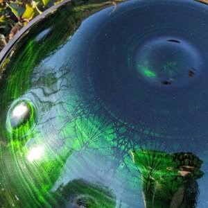 May include: Close-up of a handpan drum with a glossy, reflective surface. The instrument features a gradient of deep green and dark blue hues, with intricate patterns. The metal rim is visible, and the light reflects off the surface.