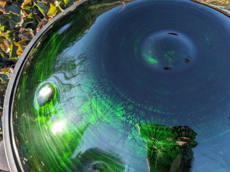 May include: Close-up of a handpan drum with a glossy, reflective surface. The instrument features a gradient of deep green and dark blue hues, with intricate patterns. The metal rim is visible, and the light reflects off the surface.