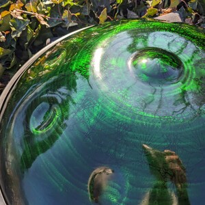 May include: A close-up of a green and blue handpan instrument. The metal surface reflects the surrounding environment, with a circular design and a central indentation. The instrument has a silver rim.