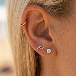 May include: A gold hoop earring in the upper ear cartilage, a small silver wave-shaped stud earring in the middle lobe, and a round, clear, faceted stud earring in the lower lobe, all worn on a single ear with blonde hair visible.