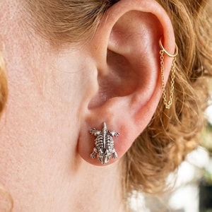 May include: A silver-toned turtle stud earring worn on the earlobe, paired with a gold-toned hoop earring featuring a delicate dangling chain that connects to the hoop, creating a layered look on the upper ear cartilage.