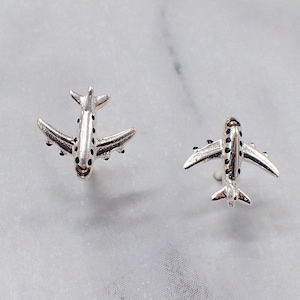 Può includere: Due orecchini a lobo a forma di aeroplano argentati. Ogni orecchino presenta un design dettagliato di aeroplano con ali e fusoliera. Gli orecchini sono esposti su una superficie di marmo bianco e grigio.