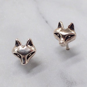May include: A pair of silver fox head stud earrings. The fox heads are facing forward with black eyes and a detailed snout.