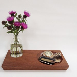 May include: A wooden tray with two compartments. One compartment holds a glass vase with a bouquet of purple thistle flowers. The other compartment holds a watch and a small pocket knife.