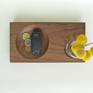 May include: A wooden tray with a round indentation for holding keys and coins. The tray also has a small vase for holding a bouquet of yellow flowers.