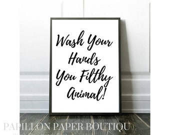 May include: A black and white printable sign with the text "Wash Your Hands You Filthy Animal!" in a cursive font. The sign is framed in a black frame and is sitting on a wooden surface.