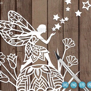 May include: White paper cut-out design of a fairy with wings, a flower, and stars. The fairy is holding a star in her hand. The design is on a wood background.