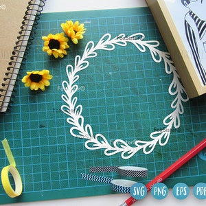 May include: A white paper wreath with a leafy design, surrounded by yellow sunflowers on a green cutting mat. The image also includes a pencil, a roll of yellow tape, and a roll of patterned tape.