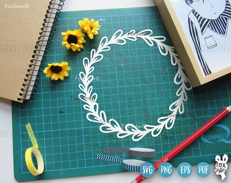May include: A white paper wreath with a leafy design, surrounded by yellow sunflowers on a green cutting mat. The image also includes a pencil, a roll of yellow tape, and a roll of patterned tape.