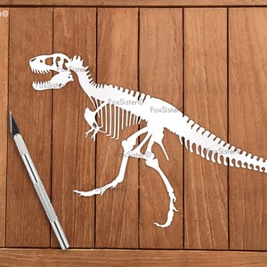K&ouml;nnte beinhalten: Wei&szlig;er Papierschnitt eines Dinosaurierskeletts auf einem Holzgrund. Ein silbernes Bastelmesser ist ebenfalls im Bild.