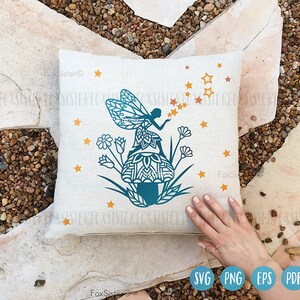 May include: A white pillow with a blue and teal silhouette of a fairy sitting on a mushroom with stars and flowers around it.