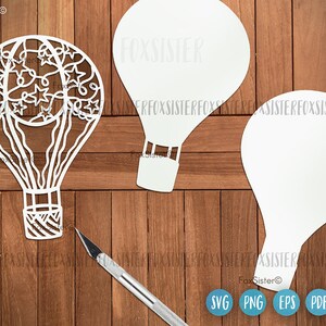 May include: Three white paper cutouts of hot air balloons. One balloon has a detailed design of stars and swirls. The other two balloons are plain. The cutouts are on a wooden surface.