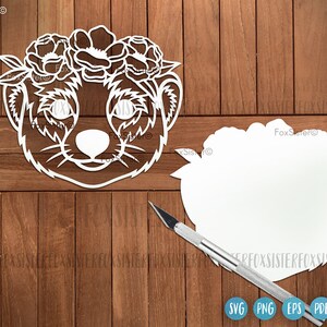 May include: White paper cut-out of a meerkat wearing a flower crown. The meerkat is facing forward with its eyes closed. The flower crown is made of simple flower shapes. The cut-out is on a wooden background.